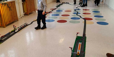 Powiększ grafikę: Rodzice , uczniowie i instruktor podczas sterowania składem pociągu z klocków na korytarzu szkolnym.