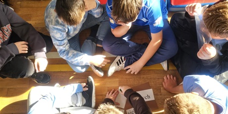 Powiększ grafikę: Uczniowie siedzą na podłodze w sali gimnastycznej, rozwiązują zadania.