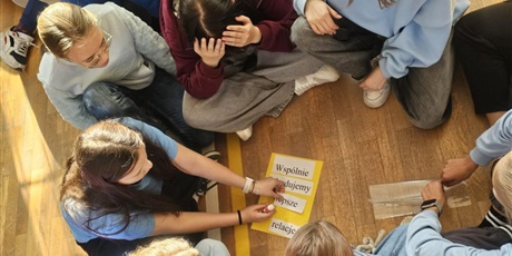 Powiększ grafikę: Uczniowie siedzą na podłodze w sali gimnastycznej, rozwiązują zadania.