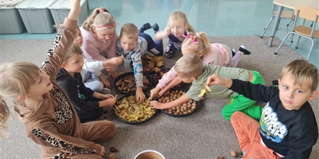 Powiększ grafikę: Uczniowie siedzą na dywanie w kole w sali lekcyjnej. Na środku misy z orzechami, szyszkami itp