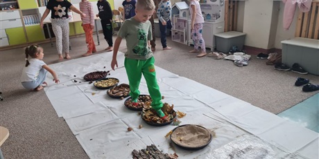 Powiększ grafikę: Na podłodze misy z różnymi nasionami ustawione w jednej linii. Uczennica pokonuje "ścieżkę sensoryczną".