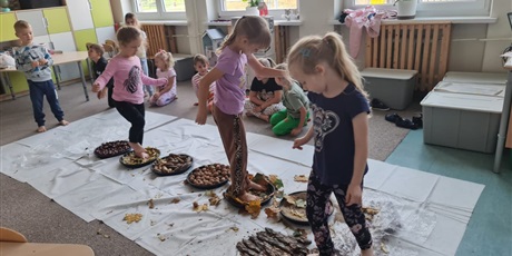 Powiększ grafikę: Na podłodze misy z różnymi nasionami ustawione w jednej linii. Uczniowie  pokonują "ścieżkę sensoryczną".