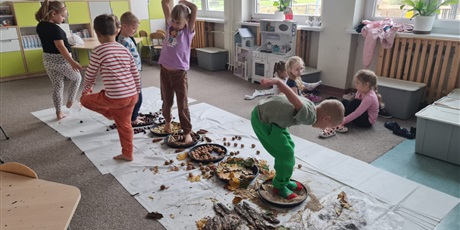 Powiększ grafikę: Na podłodze misy z różnymi nasionami ustawione w jednej linii. Uczniowie  pokonują "ścieżkę sensoryczną".