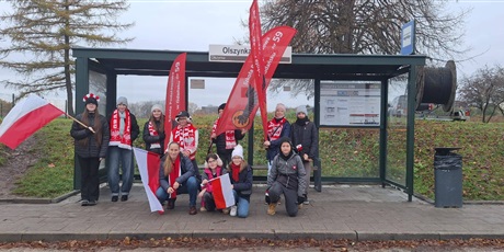 Powiększ grafikę: Uczniowie z flagami, symbolami narodowymi  i banerami szkolnymi na przystanku autobusowym w drodze na paradę niepodległości.