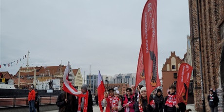 Powiększ grafikę: Uczniowie z flagami, symbolami narodowymi  i banerami szkolnymi na ulicy w drodze na paradę niepodległości.