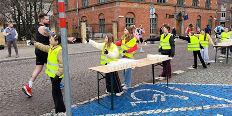 Powiększ grafikę: Biegacze oraz wolontariusze podczas podawania napojów na trasie maratonu.